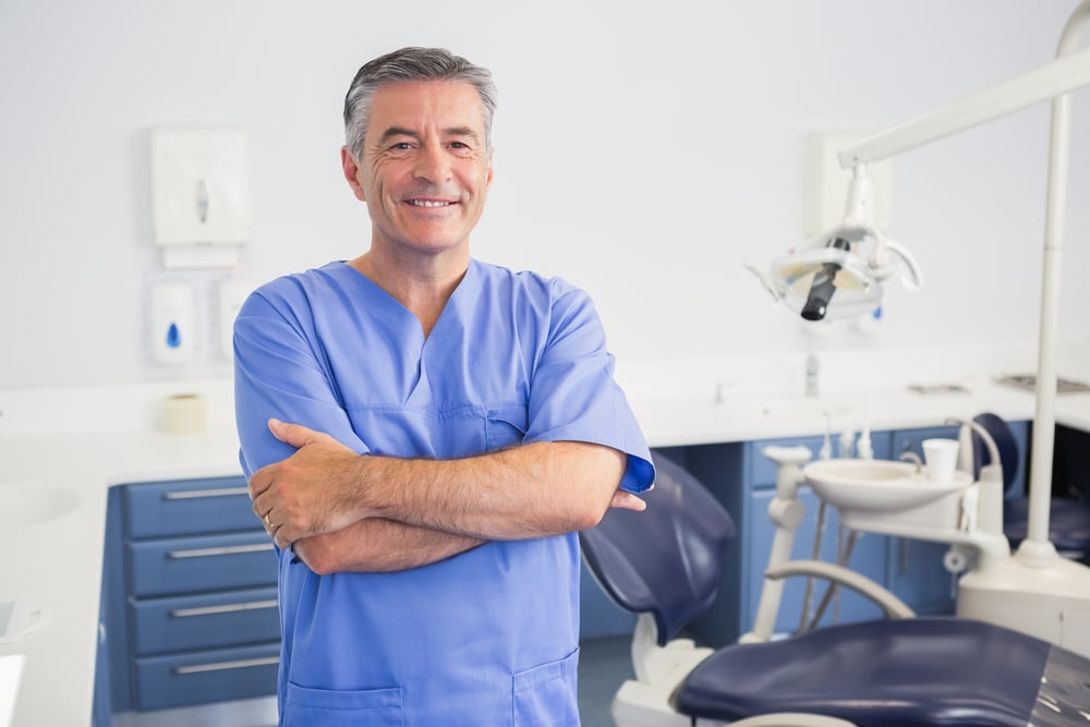 A smiling male dentist standing in his office with his arms crossed